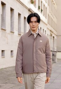 Padded, light brown jacket with a collar, front button closure, and a logo patch on the chest. Worn with a beige shirt and trousers.