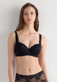 Czarny koronkowy biustonosz z usztywnianymi miseczkami, falistym wykończeniem i regulowanymi ramiączkami. Ozdobiony kwiatowymi wzorami i gładkim pasem pod biustem zapewniającym wsparcie.