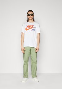 Hvid t-shirt med et rødt og orange Nike-logo, parret med lysegrønne bukser og hvide sneakers. Tilbehøret inkluderer mørke solbriller.