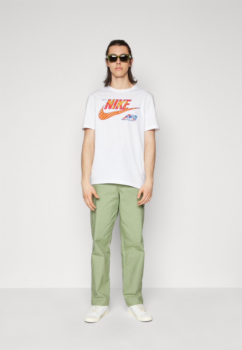 T-shirt blanc avec un logo Nike rouge et orange, associé à un pantalon vert clair et des baskets blanches. Les accessoires incluent des lunettes de soleil noires.