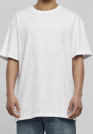 Osoba nosząca prosty, bardzo luźny biały t-shirt z krótkim rękawem i niebieskie dżinsy, stojąca na szarym tle.