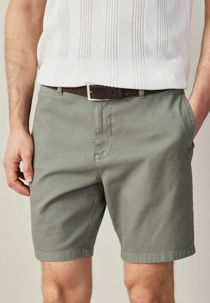 Man wearing olive green shorts and a white textured sleeveless top, with a dark brown belt and hands in pockets.