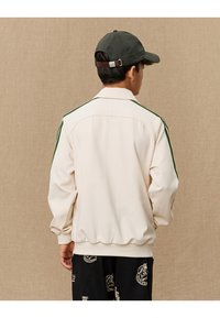 Lys beige jakke med grønne sidestriper, ribbestrikkede mansjetter og hemming. Glatt tekstur, avslappet passform. Brukt med svarte mønstrede bukser og caps.