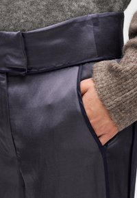 Χέρι στην τσέπη σκούρων μπλε, γυαλιστερών παντελονιών που φοριούνται με καφέ πλεκτό πουλόβερ.