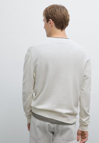 Cremfarbener Strickpullover mit Rundhalsausschnitt, der eine strukturierte Oberfläche und gerippte Manschetten aufweist. Kombiniert mit hellgrauen Hosen.