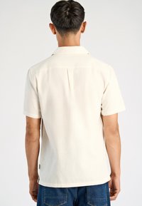 Camisa de manga corta, de color crema claro, hecha de una tela texturizada. Presenta un cuello clásico y un detalle de abertura en la parte posterior, combinada con jeans azules.