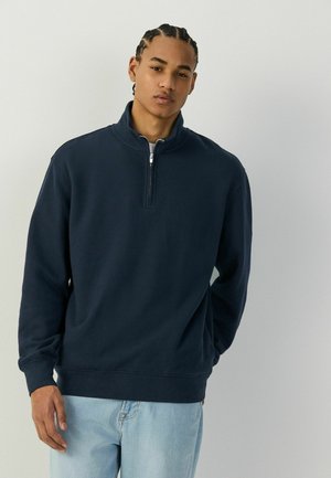 Young man with braided hair wearing a navy blue quarter-zip sweatshirt and light blue jeans, standing against a plain light background.
