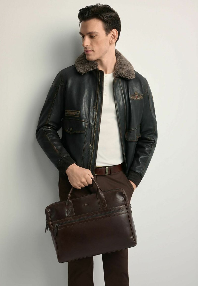 Brązowa skórzana torba na dokumenty z dwoma górnymi uchwytami i przednią kieszenią na zamek, w zestawieniu z czarną skórzaną kurtką z futrzanym kołnierzem i złotymi akcentami.