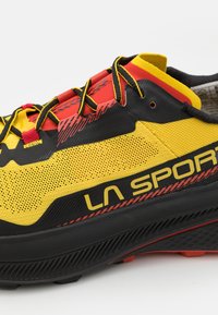 Scarpa sportiva nera e gialla con accenti rossi. Presenta materiale in rete, suola spessa, branding prominente e elementi di design testurizzati.