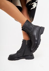 Botas de cuero negras hasta el tobillo con cordones en la parte delantera, cremallera lateral y suela de goma gruesa. Cuero texturizado con acentos lisos, punta redondeada.
