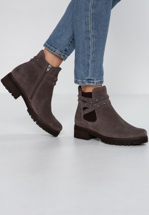 Bottines en daim marron avec fermeture éclair latérale et lanières décoratives cloutées portées avec un jean en denim bleu.