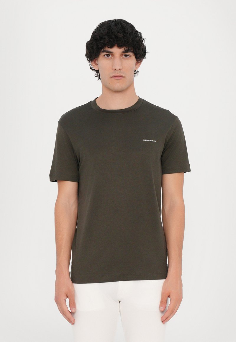 T-shirt vert olive à manches courtes en coton, avec col ronde et un petit logo blanc sur la poitrine. Texture lisse.
