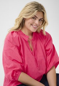 Blusa rosa con scollo arricciato e dettaglio con cordoncino. Presenta maniche a palloncino e un motivo ricamato su tutta la superficie.