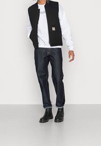 Gilet senza maniche nero sopra un felpa bianca, abbinato a jeans blu scuro. Presenta una tasca frontale e un collo rotondo. Stivali neri alla caviglia.