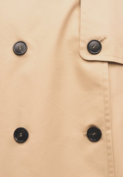 Μπεζ ύφασμα με τέσσερα μαύρα κουμπιά τοποθετημένα σε δύο κάθετα ζευγάρια, εμφανίζοντας λεπτομέρειες ραφής στο ένδυμα.