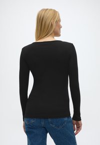 Γυναίκα με ξανθά μαλλιά που φοράει εφαρμοστή μαύρη μπλούζα με μακριά μανίκια και μπλε τζιν, με την πλάτη προς τα έξω σε απλό λευκό φόντο.