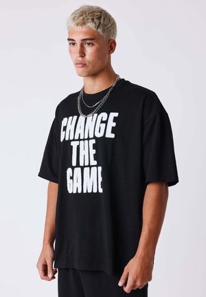 Young man with short blond hair wears a black oversized T-shirt with bold white text "CHANGE THE GAME" and two silver chain necklaces.