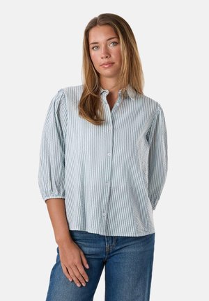 Frau mit hellbraunen Haaren, die eine hellblau-weiß gestreifte Hemdbluse und blaue Jeans trägt, steht vor einem schlichten Hintergrund.