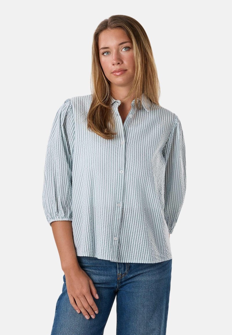 Femme aux cheveux châtain clair portant une chemise boutonnée à rayures bleu clair et blanc et un jean bleu, se tenant devant un fond uni.