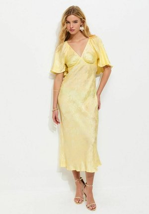 Vrouw draagt een lichtgele midi-jurk met korte mouwen, V-hals en bloemenpatroon, gecombineerd met gouden hooggehakte sandalen, staand tegen een witte muur.