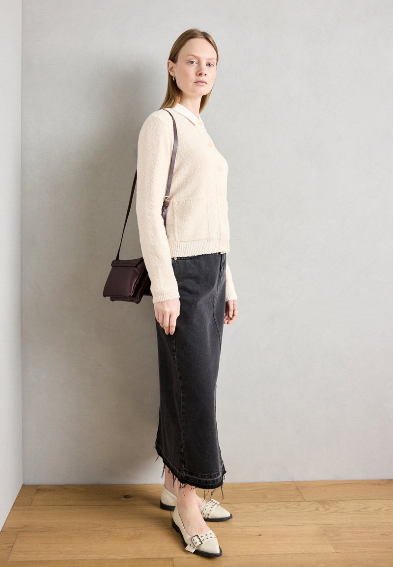 Helle beige Strickjacke mit Knöpfen aus strukturiertem Material, kombiniert mit einem schwarzen Jeansrock mit fransigem Saum und cremefarbenen Ballerinas mit Nieten.