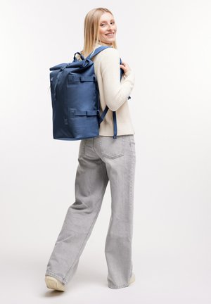 Lächelnde Frau trägt cremefarbenen Pullover und graue Jeans, mit großem blauen Roll-Top-Rucksack, blickt über die Schulter vor weißem Hintergrund.