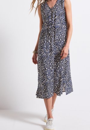 Femme portant une robe midi sans manches à motif bleu et blanc avec un design portefeuille et des baskets blanches, debout devant un fond clair uni.