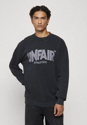 Jeune homme aux cheveux bouclés porte un sweat-shirt noir avec "UNFAIR ATHLETICS" imprimé sur le devant, debout devant un fond uni.