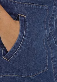 Denim oblačilo s bližinskim posnetkom ukrivljenega žepa. Tkanina je temno modra, z kontrastnim šivanjem in gladko teksturo.
