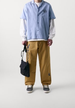 Light blue short-sleeve shirt with logo, white long-sleeve shirt underneath, tan pants with embroidered designs, black bag, gray sneakers.