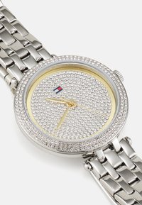 Reloj de metal plateado con una caja redonda, esfera engastada de cristal, acentos en oro amarillo y una pulsera de metal con eslabones. Presenta tres agujas doradas.
