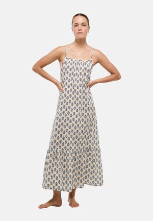 Femme debout pieds nus, mains sur les hanches, portant une robe maxi blanche sans manches avec un motif floral noir sur un fond uni.