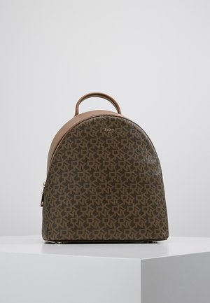 Compact bruine DKNY-rugzak met monogrampatroon, gebogen bovenhandgreep en gouden rits, tentoongesteld op een wit voetstuk tegen een grijze achtergrond.