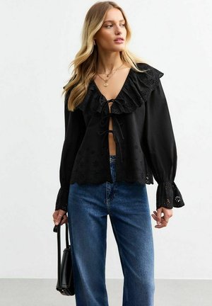 Black long-sleeve blouse with a ruffled collar, lace detailing, and a front tie closure; paired with blue high-waisted jeans.