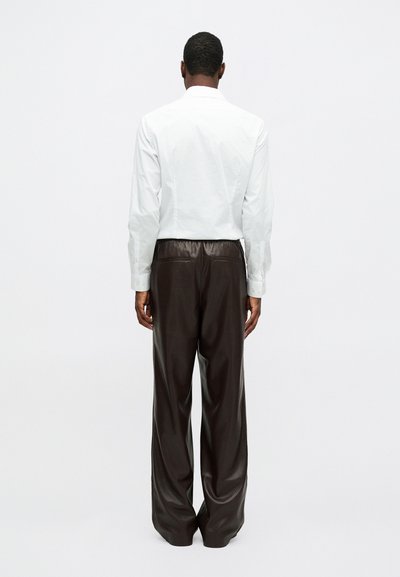 Moški obrnjen stran, oblečen v belo majico z dolgimi rokavi in ohlapne temno rjave usnjene hlače pred enobarvno svetlo ozadje.