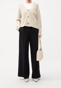 Beige gestrickte Strickjacke mit goldenen Knöpfen, kombiniert mit schwarzen, weit geschnittenen Hosen. Accessoires: kleine beige Handtasche und cremefarbene, spitz zulaufende High Heels.