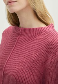 Maglione rosa lavorato a maglia con collo alto e texture a coste. Presenta un sottile dettaglio di cucitura lungo la spalla e il corpo.