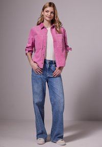 Rosa kordt skjorta, knäppning framtill, två bröstfickor; kombinerad med ljusblå vida jeans; kvinna som bär en vit t-shirt och avslappnade skor.