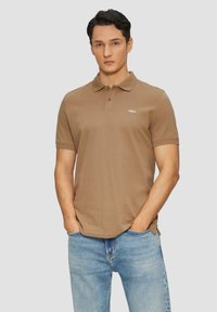 Camisa polo marrón con cuello clásico, mangas cortas y un pequeño bordado de logo en el pecho. Combinada con unos jeans azul claro.