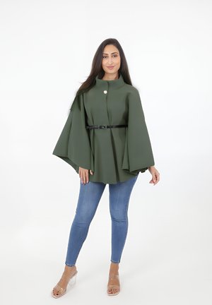 Grøn kapjakke i tekstureret stof med høj krave, frontknap og en sort bælte i taljen, parret med blå skinny jeans og gennemsigtige hæle.