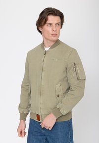 Jonge man in een lichtgroene bomberjas met een ritssluiting op de mouwzak en blauwe spijkerbroek, kijkend naar links tegen een effen achtergrond.