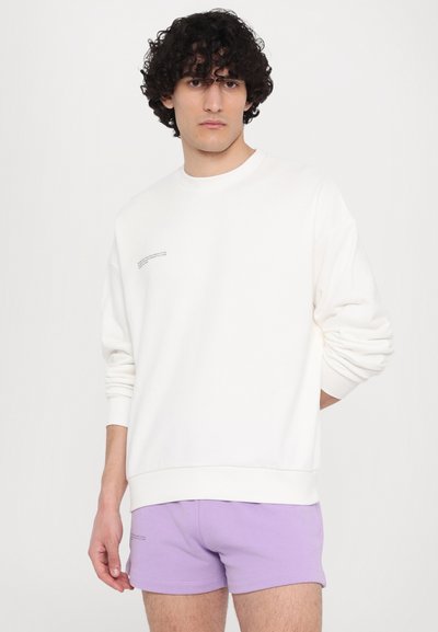 Hvid sweatshirt med rund hals og ribbede manchetter, parret med lilla shorts. Bemærkelsesværdige detaljer inkluderer tekst på sweatshirtens forside.