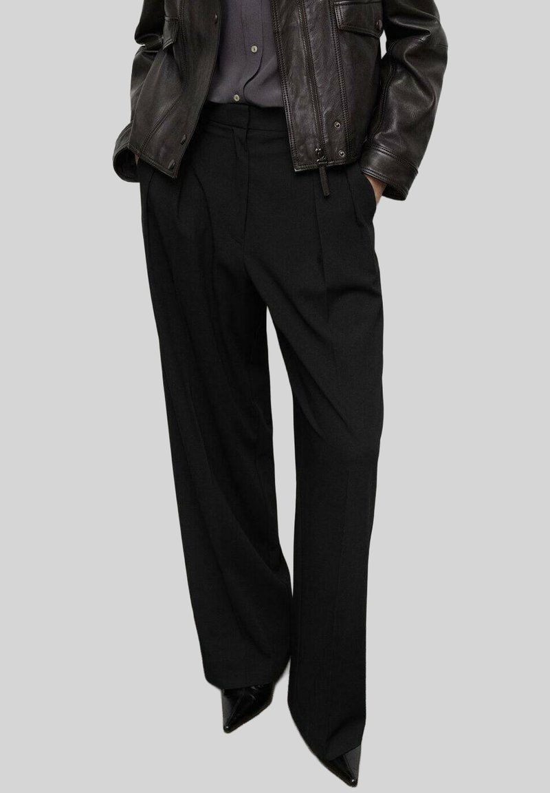 Czarne szerokie spodnie o dopasowanym kroju, z plisami, wysokim stanem i gładką fakturą, w połączeniu z czarnymi, spiczastymi butami.