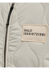 Quilted light beige jacket featuring a zip closure, black zipper, and a rectangular tag with "HALO 196819721981" in bold black text.