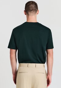 Uomo con una camicia a maniche corte verde scuro infilata in pantaloni beige, in piedi di spalle contro uno sfondo chiaro e uniforme.