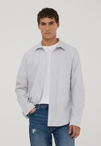 Camicia a righe blu chiaro e bianca, maniche lunghe, taschino sul petto, abbinata a una maglietta bianca e jeans blu.