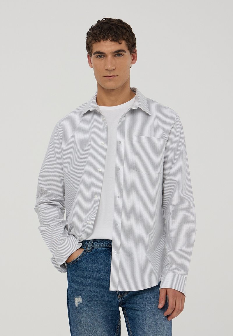 Camicia a righe blu chiaro e bianca, maniche lunghe, taschino sul petto, abbinata a una maglietta bianca e jeans blu.