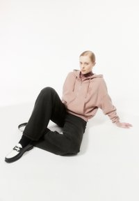 Haut de sweatshirt zippé rose clair avec une poche avant et un logo, associé à un pantalon large noir et des mocassins blancs avec une sangle noire. Texture de tissu lisse.