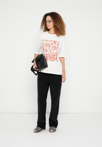 Λευκό κοντομάνικο μπλουζάκι με κόκκινο γραφικό σχέδιο που απεικονίζει φοίνικες και κείμενο, σε συνδυασμό με μαύρο παντελόνι άνετης γραμμής και μαύρη τσάντα.