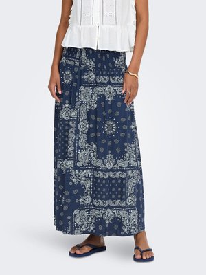 Femme portant une jupe longue bleu marine à motifs paisley blancs, un top blanc sans manches, un bracelet en or, une bague et des tongs bleu marine.